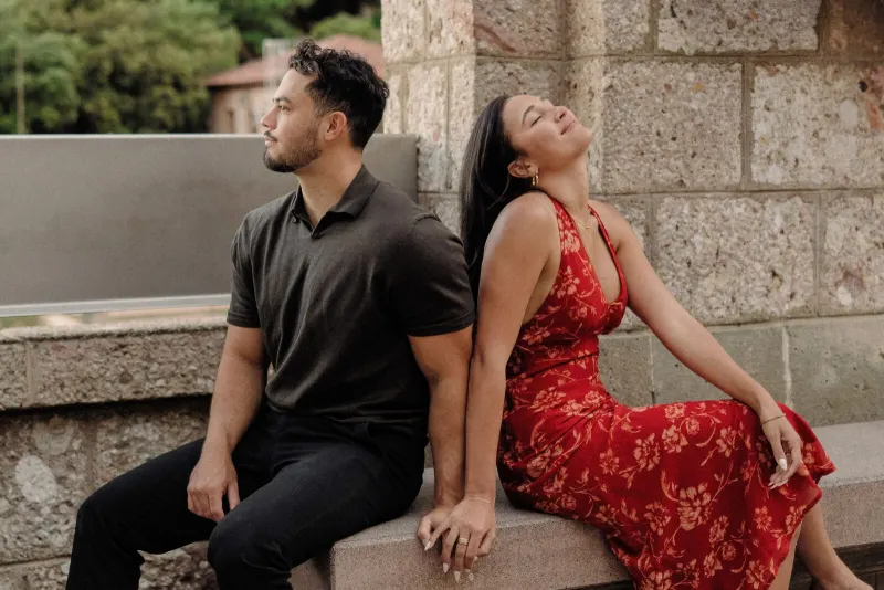 Dan and Sydney sit back to back beside a stone wall during their Montserrat engagement session.