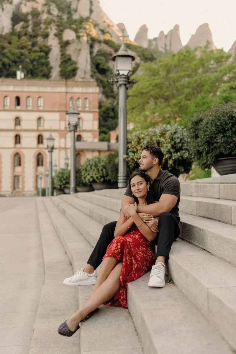 Dan and Sydney sit together on stone steps with Montserrat monastery and mountains behind them.