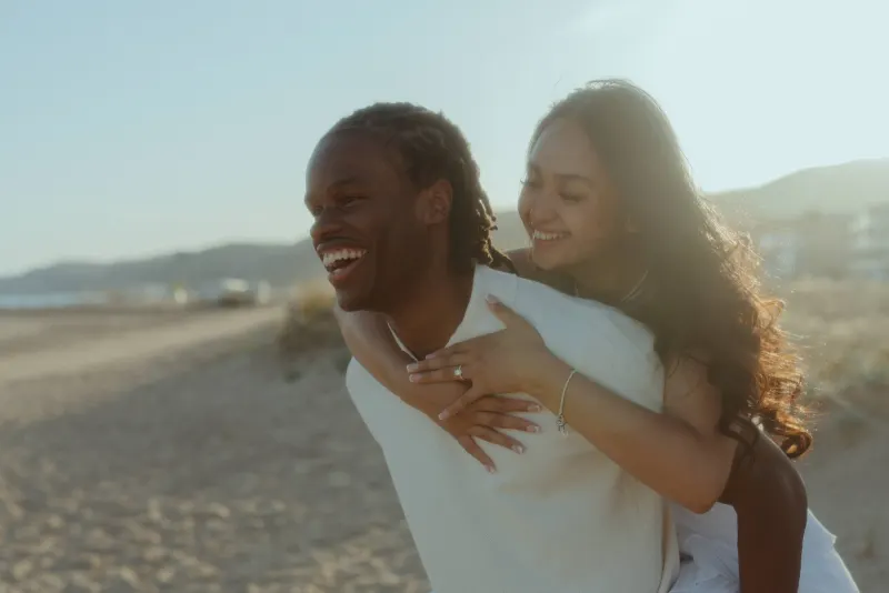 Dexter carries Cheryl on his back as they laugh during their Castelldefels Beach engagement session.