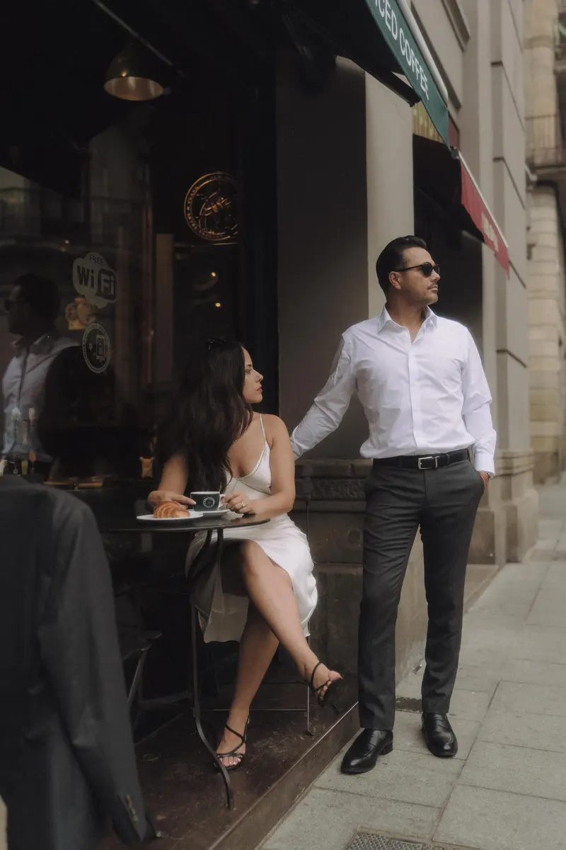 Edgar stands beside Ashley as she sits at an outdoor cafe table in Barcelona.