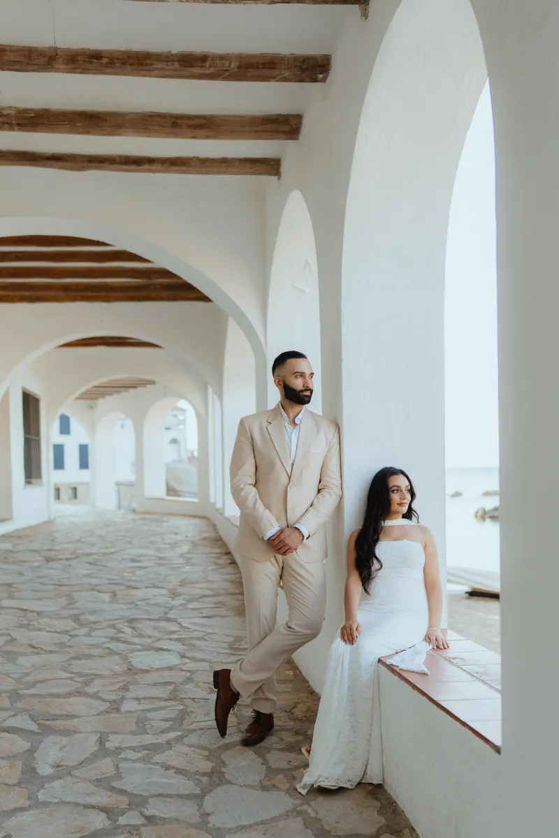 Elegant couple during a surprise proposal photoshoot, sharing a quiet moment under white arches by the sea.