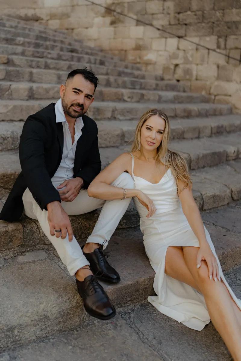 Elegant couple in white and black outfits sit close together on stone steps during a couples & engagements photoshoot.