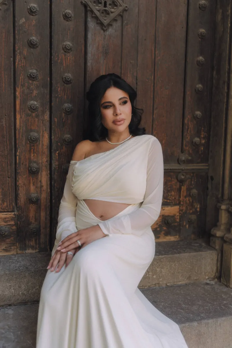 Elegant expectant mother in a draped white dress poses by a rustic wooden door during a Vacation Portraits photoshoot.