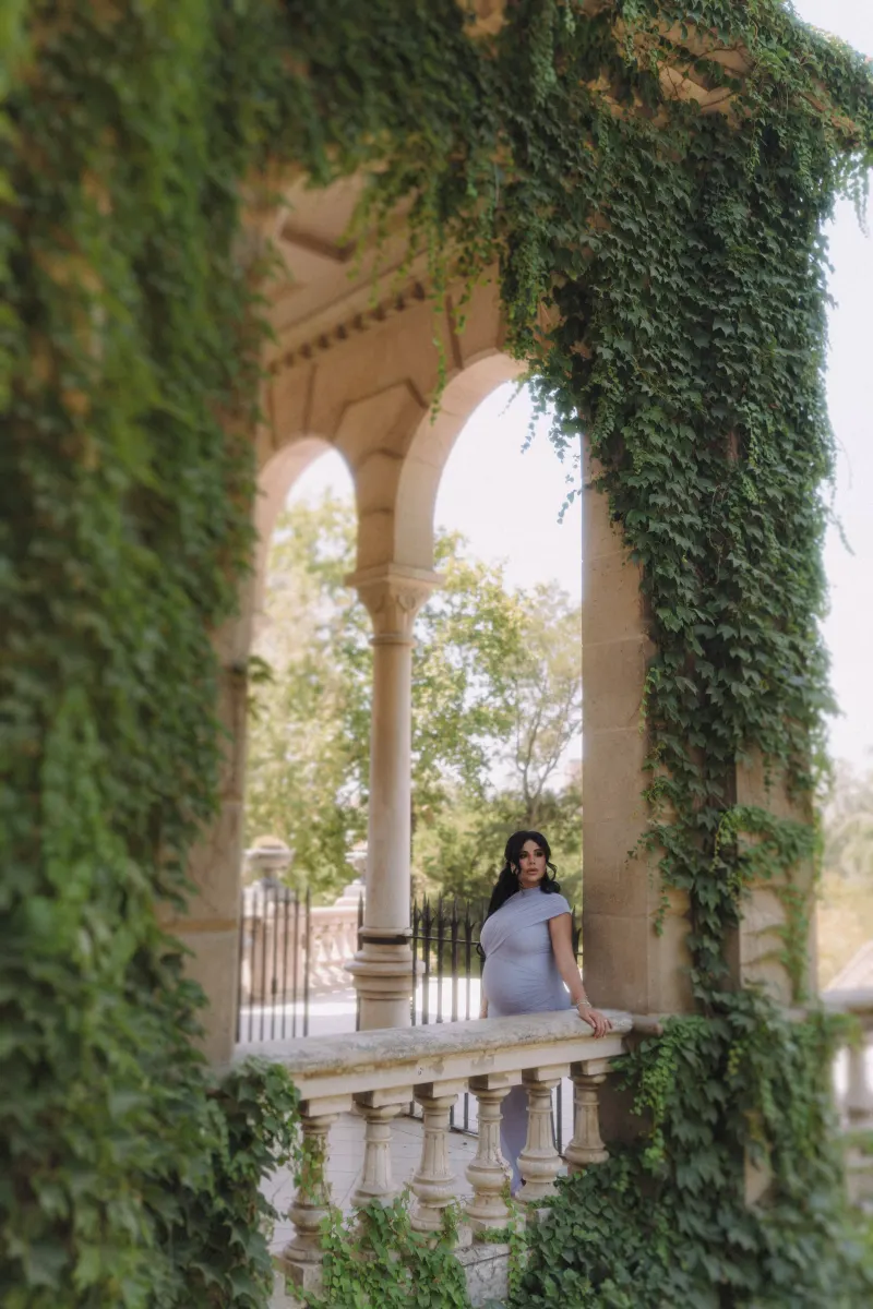 Elegant expectant mother in a flowing dress stands on a leafy balcony, Vacation Portraits session in Barcelona.