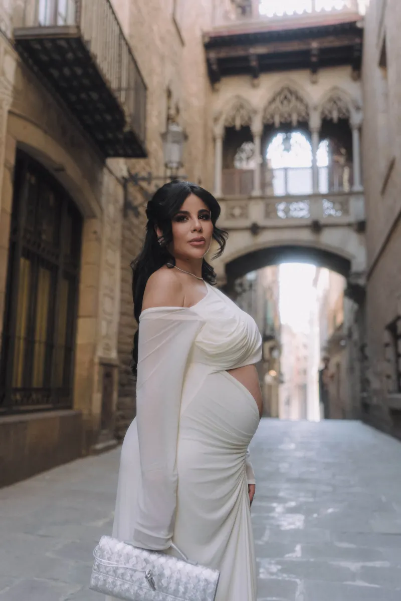 Elegant expectant mother in a flowing white dress poses confidently in a historic city alley during a Vacation Portraits shoo