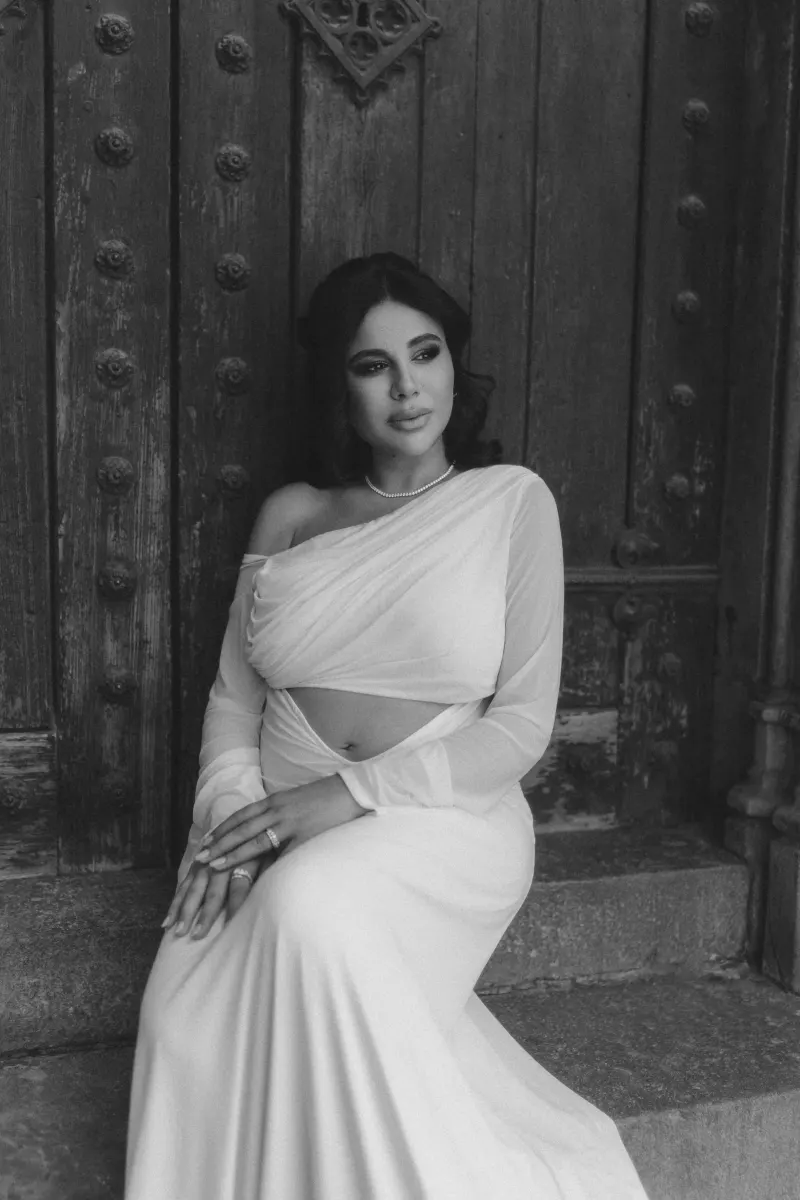 Elegant expectant mother in a white dress poses thoughtfully by a rustic wooden door during a Vacation Portraits session.