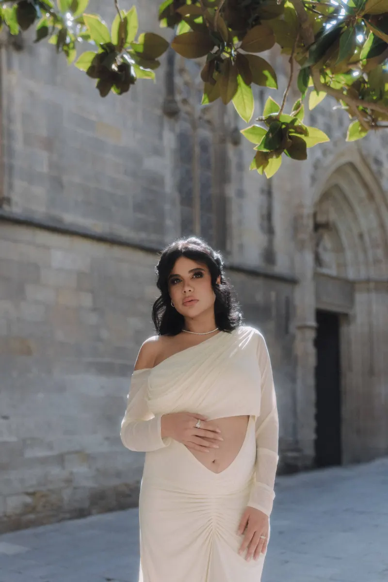 Elegant expectant mother in a white dress stands beneath leafy branches during a Vacation Portraits session in the city.