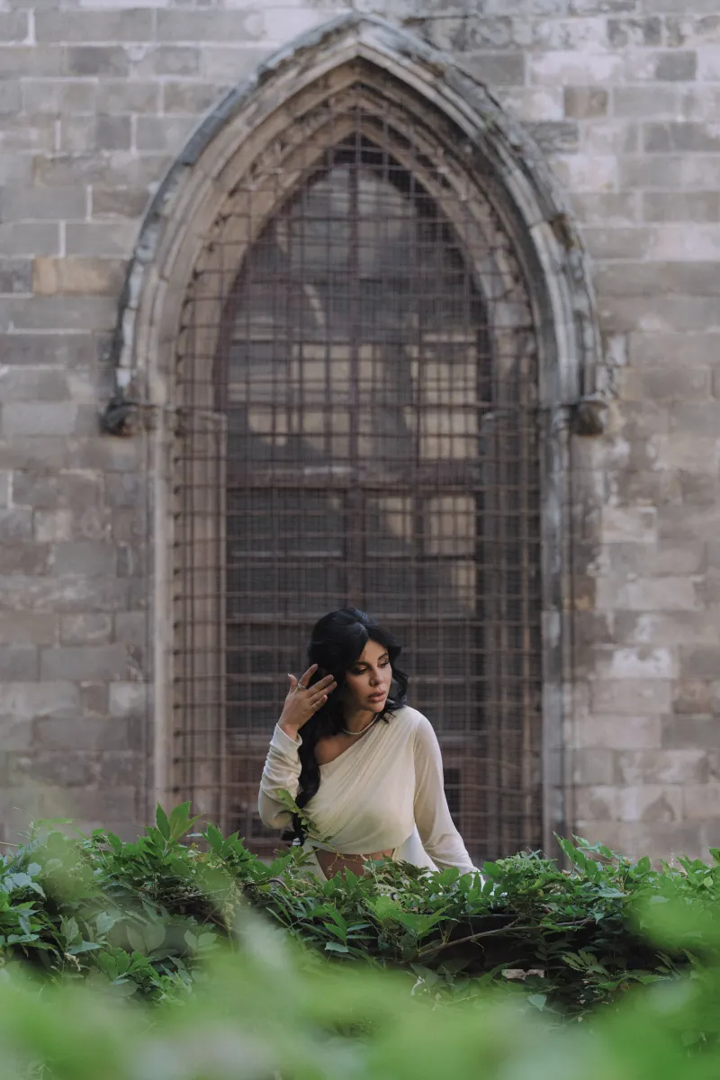 Elegant expectant mother in a white dress touches her hair beside lush greenery during a Vacation Portraits photoshoot.