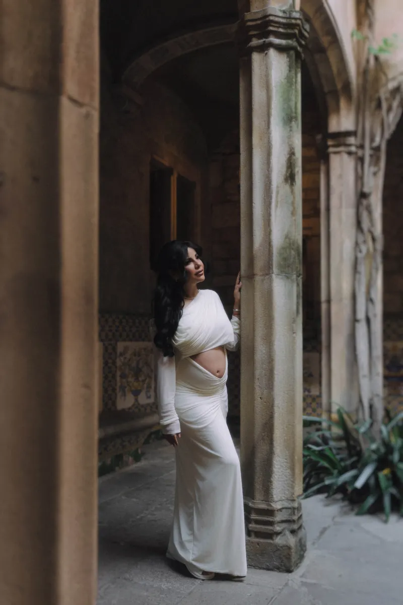 Elegant pregnant woman in a flowing white dress stands by a stone column, captured during a Vacation Portraits session.