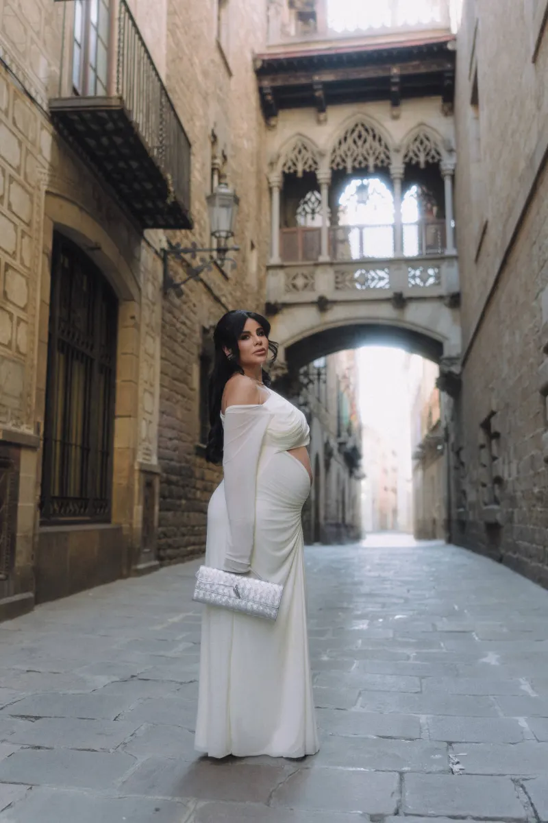 Elegant pregnant woman in a white dress poses on a quiet historic street during a Vacation Portraits photoshoot.