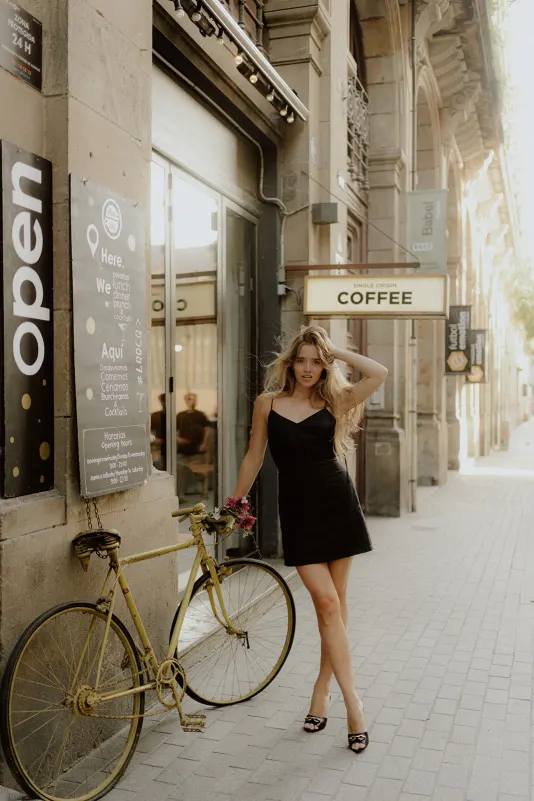 Elegant woman in a black dress poses by a yellow bike outside a café during a Vacation Portraits session in Europe.