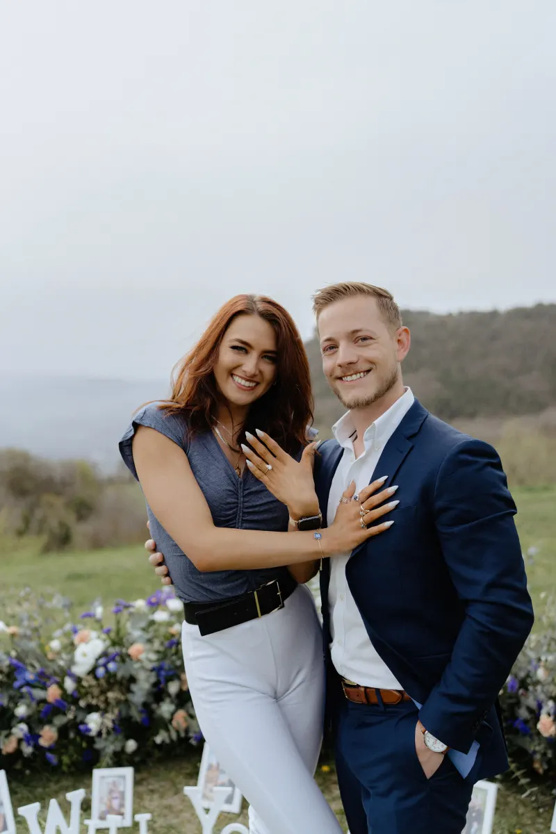 Engaged couple beams with joy, showing off rings after a countryside surprise proposal photoshoot.