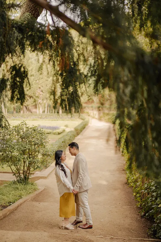 Engaged couple holding hands and smiling at each other on a garden path, couples photoshoot in a serene outdoor setting.