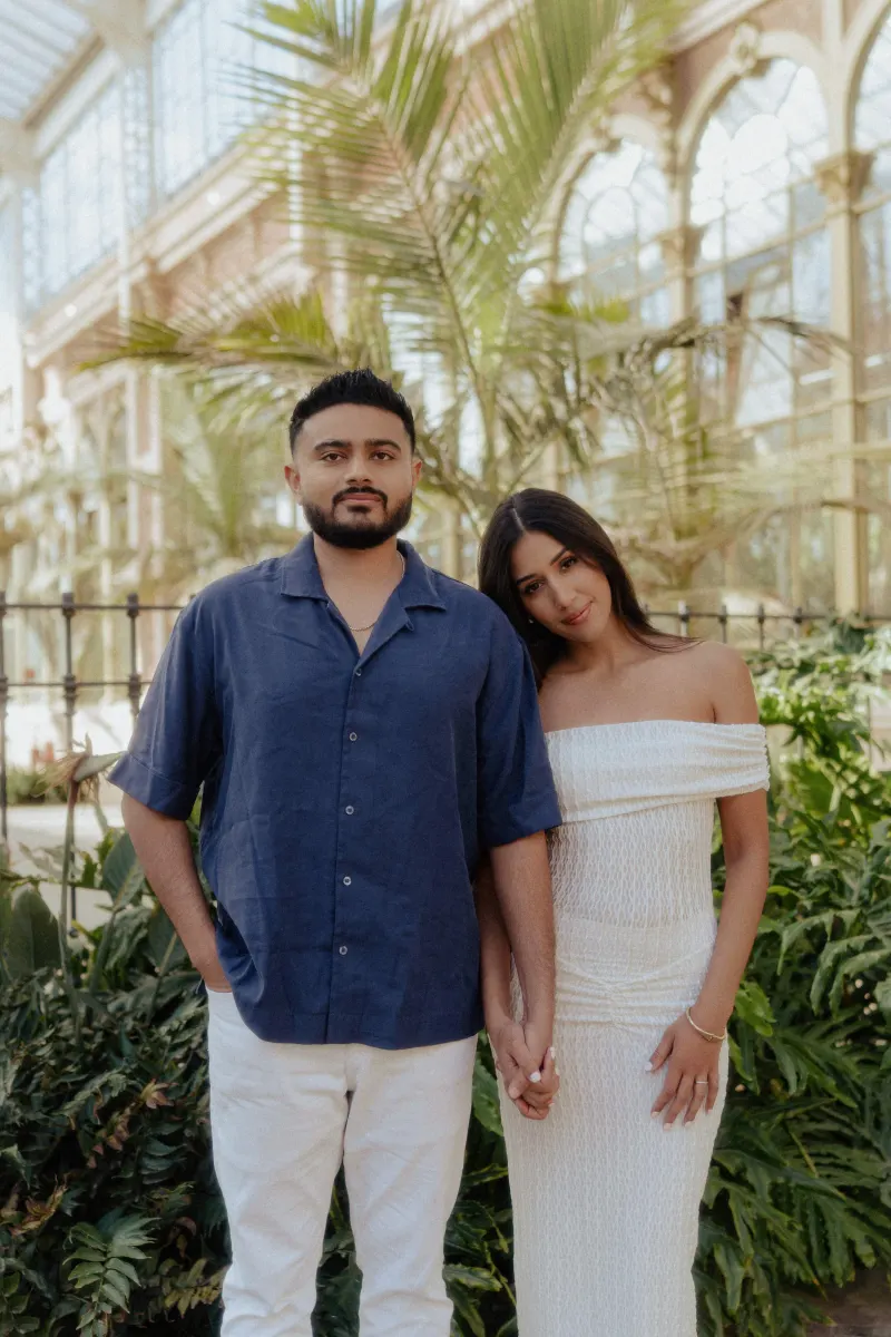 Engaged couple holding hands and smiling softly during a couples photoshoot in a bright, elegant conservatory.