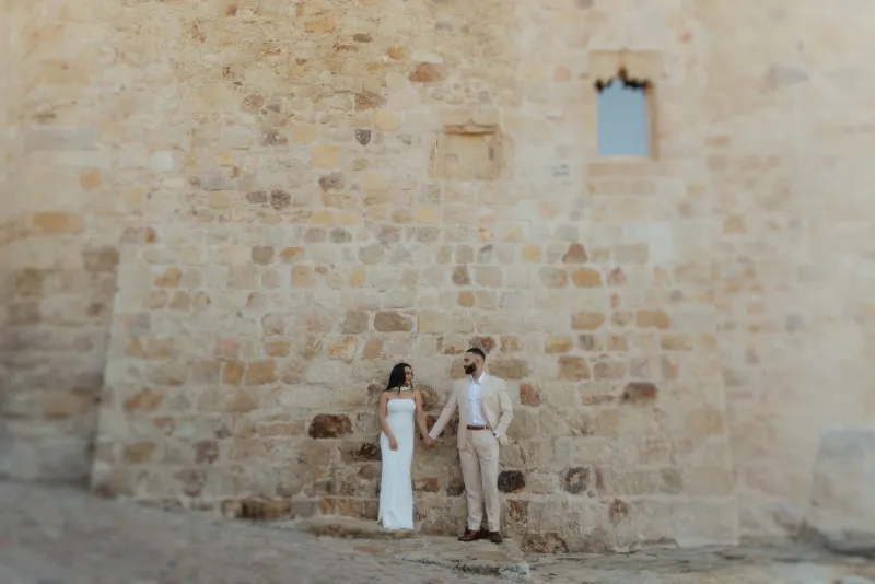 Engaged couple holds hands and smiles against a rustic stone wall, captured during a surprise proposal photoshoot.