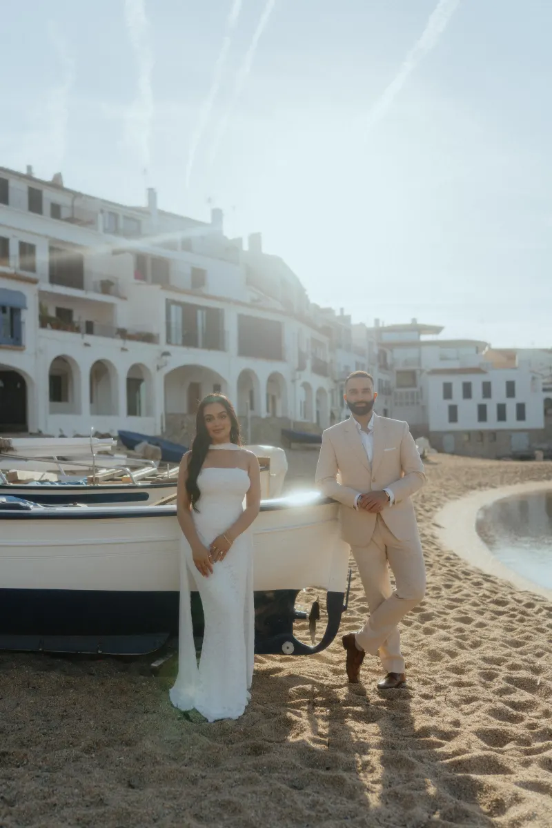Engaged couple poses by a boat on a sunlit beach in Spain during a surprise proposal photoshoot.