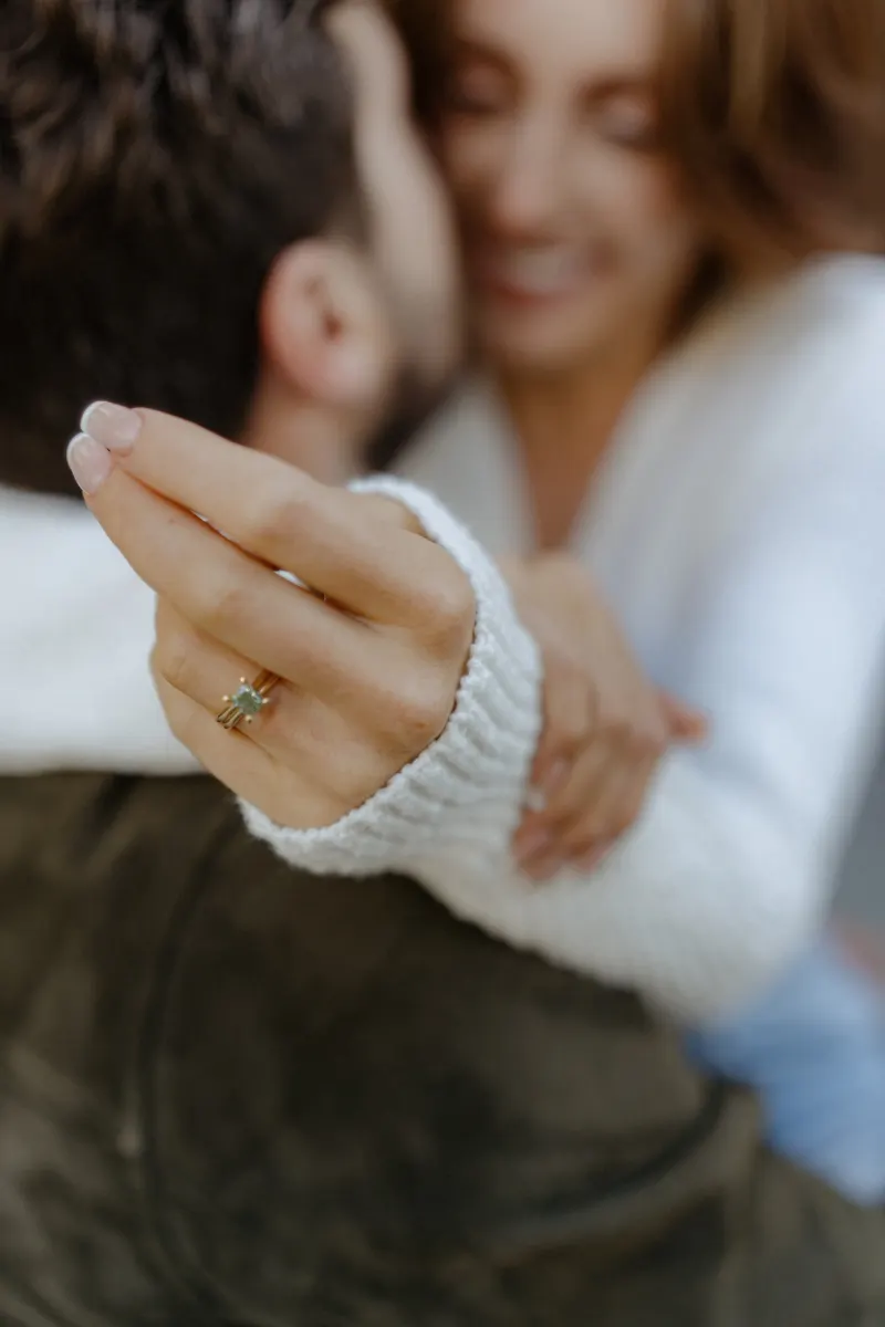Engaged couple shares a joyful embrace, her hand with an emerald ring in focus during a couples photoshoot.