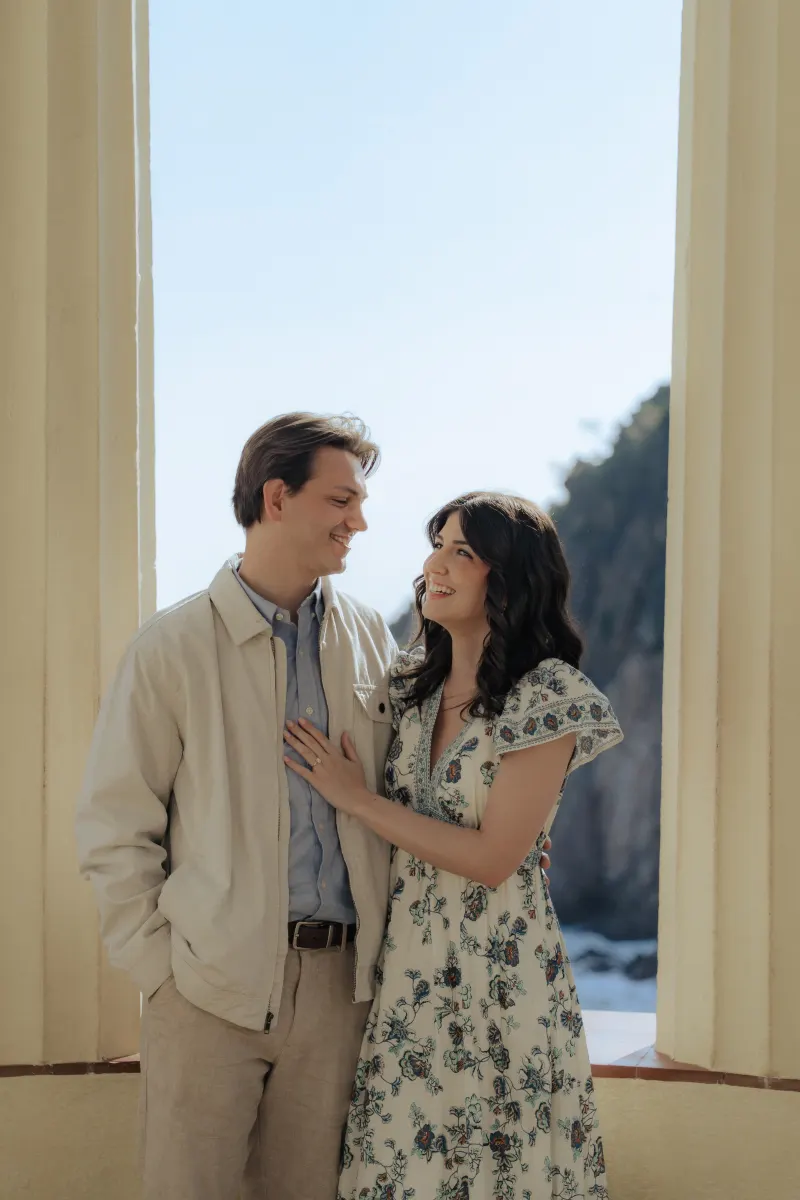 Engaged couple shares a joyful moment by the seaside columns during a surprise proposal photoshoot.