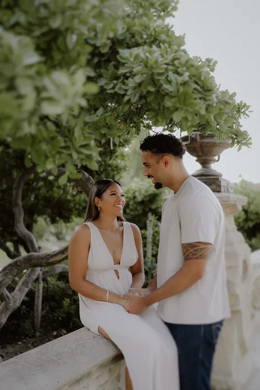 Engaged couple shares a loving gaze and holds hands beneath lush greenery during a romantic couples photoshoot.