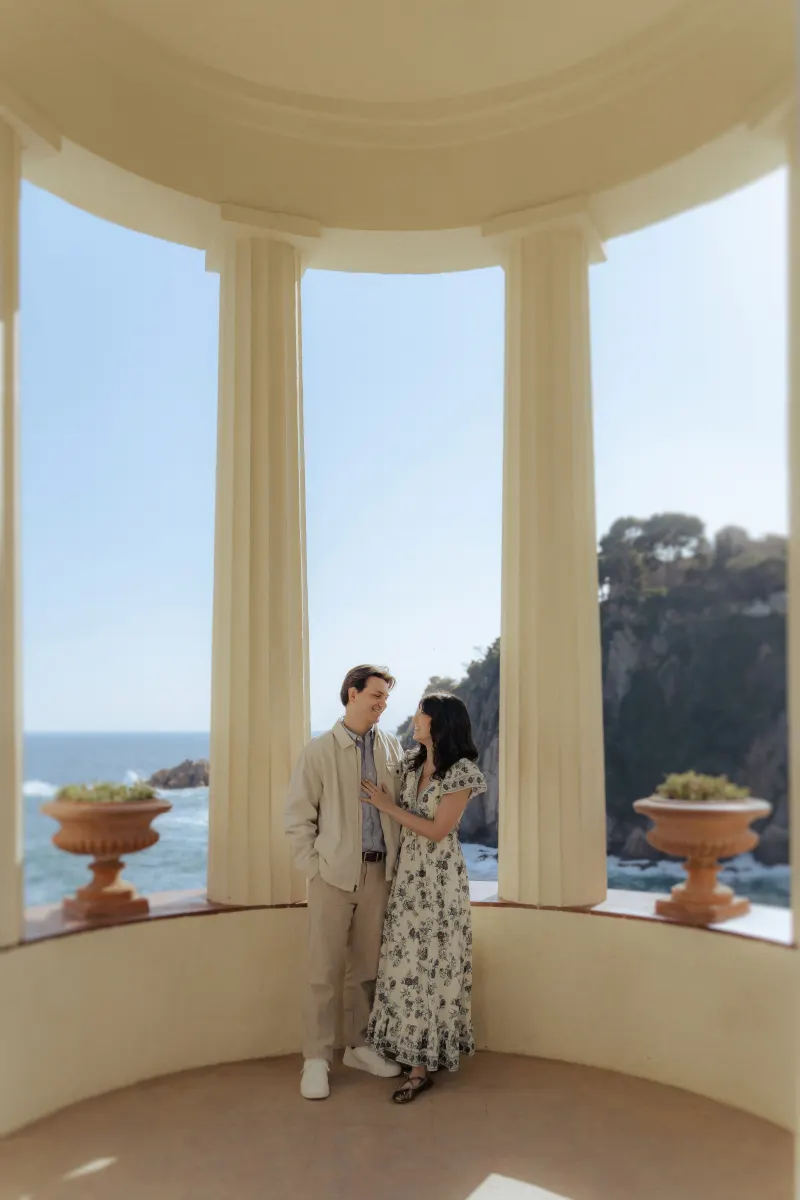 Engaged couple shares a quiet, joyful moment by the sea under elegant columns during a surprise proposal photoshoot.