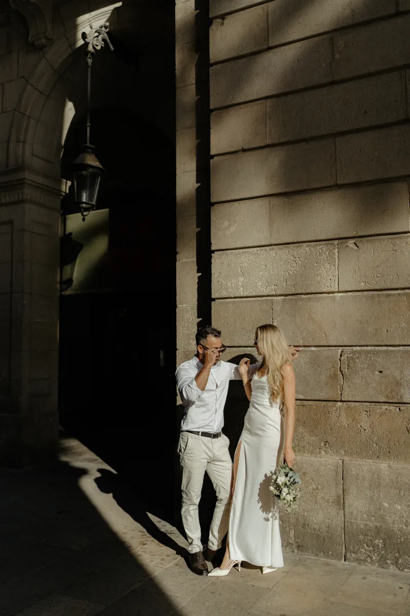 Engaged couple sharing a candid moment in elegant attire, leaning against a sunlit stone wall during a city photoshoot.