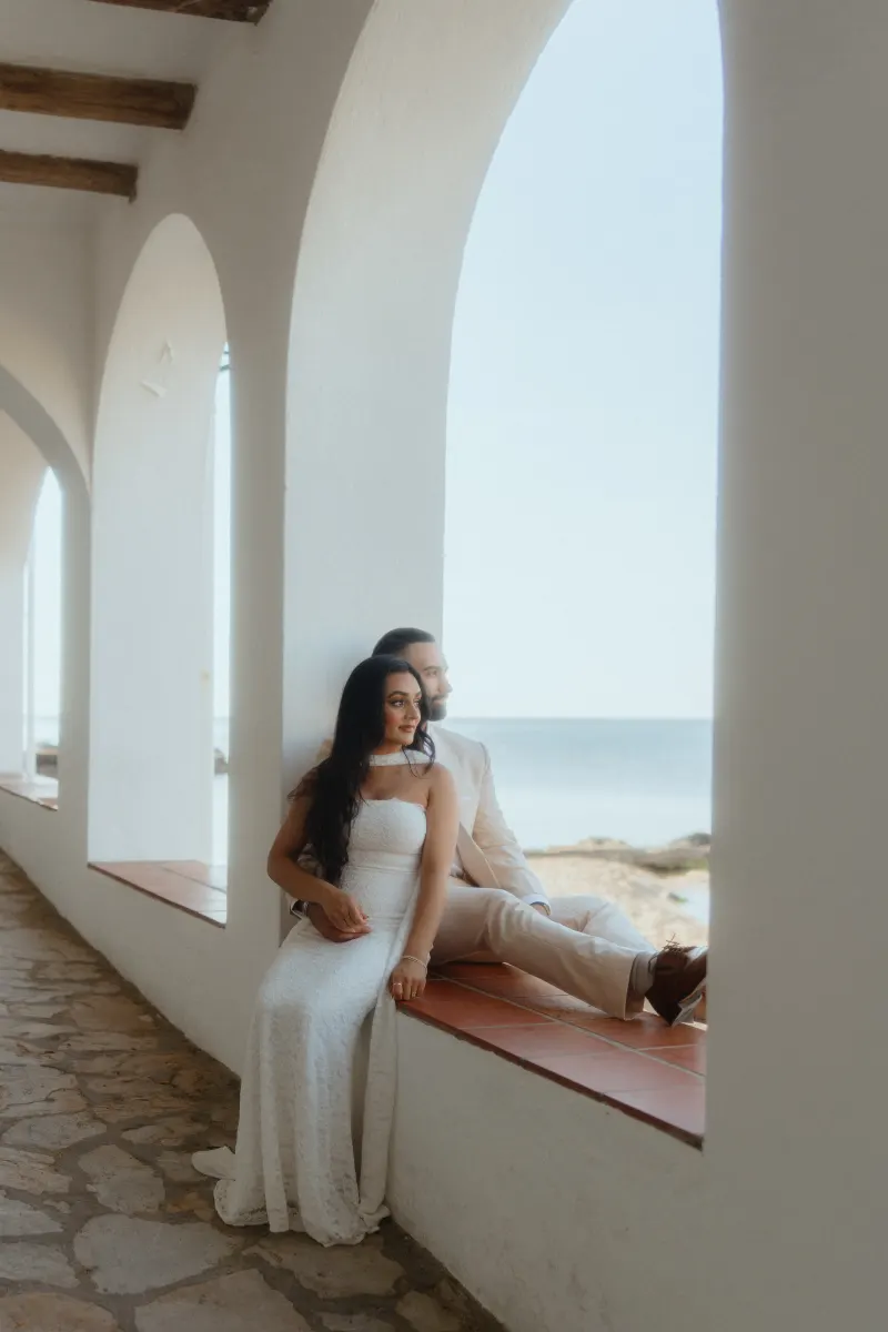Engaged couple sits in a sunlit archway by the sea, sharing a quiet moment after a surprise proposal photoshoot.