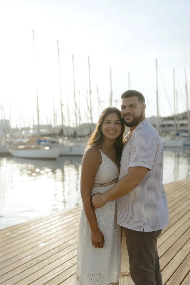 Engaged couple smiles warmly while embracing on a sunlit marina dock during a couples & engagements photoshoot.