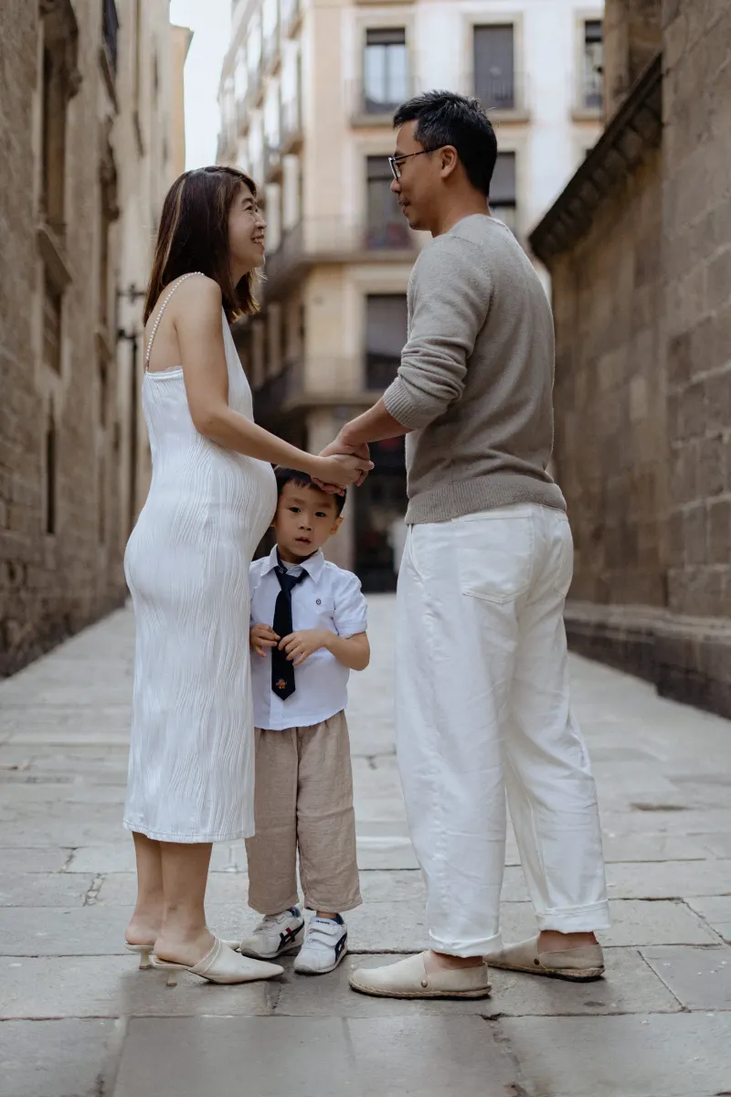 Expectant couple holding hands and smiling as their young son stands between them during a Vacation Portraits shoot in Barcel