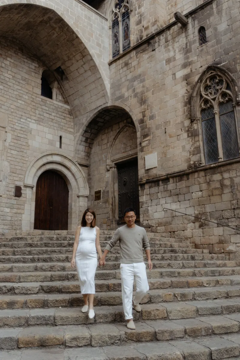 Expectant couple holding hands and walking down historic stone steps during a Vacation Portraits shoot in Barcelona.