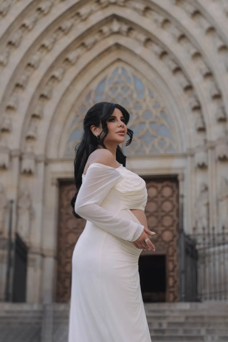 Expectant mother in a flowing white dress smiles softly during a Vacation Portraits session by a grand cathedral entrance.