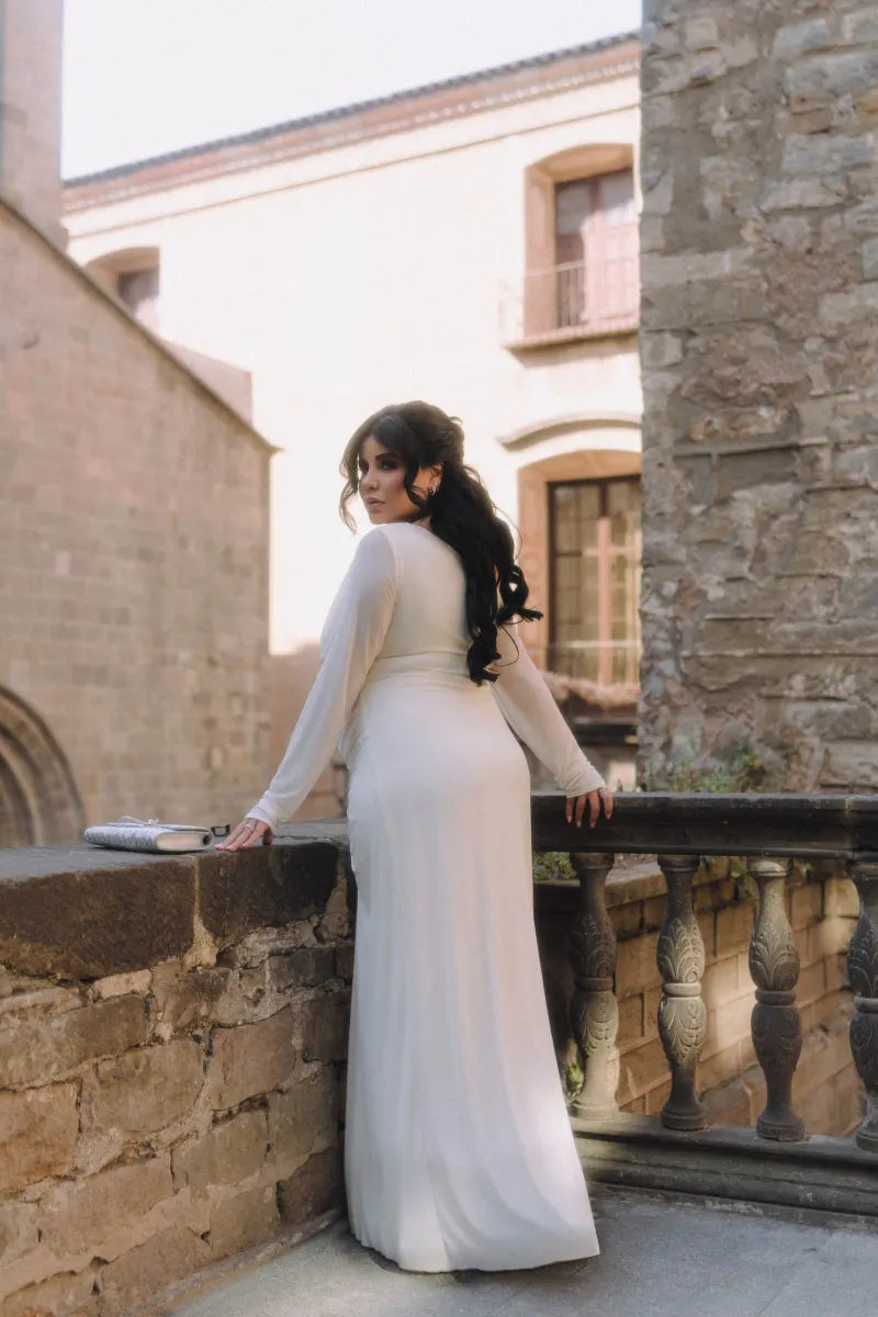 Expectant mother in a long white dress gazes back while leaning on a stone balcony during a Vacation Portraits session.