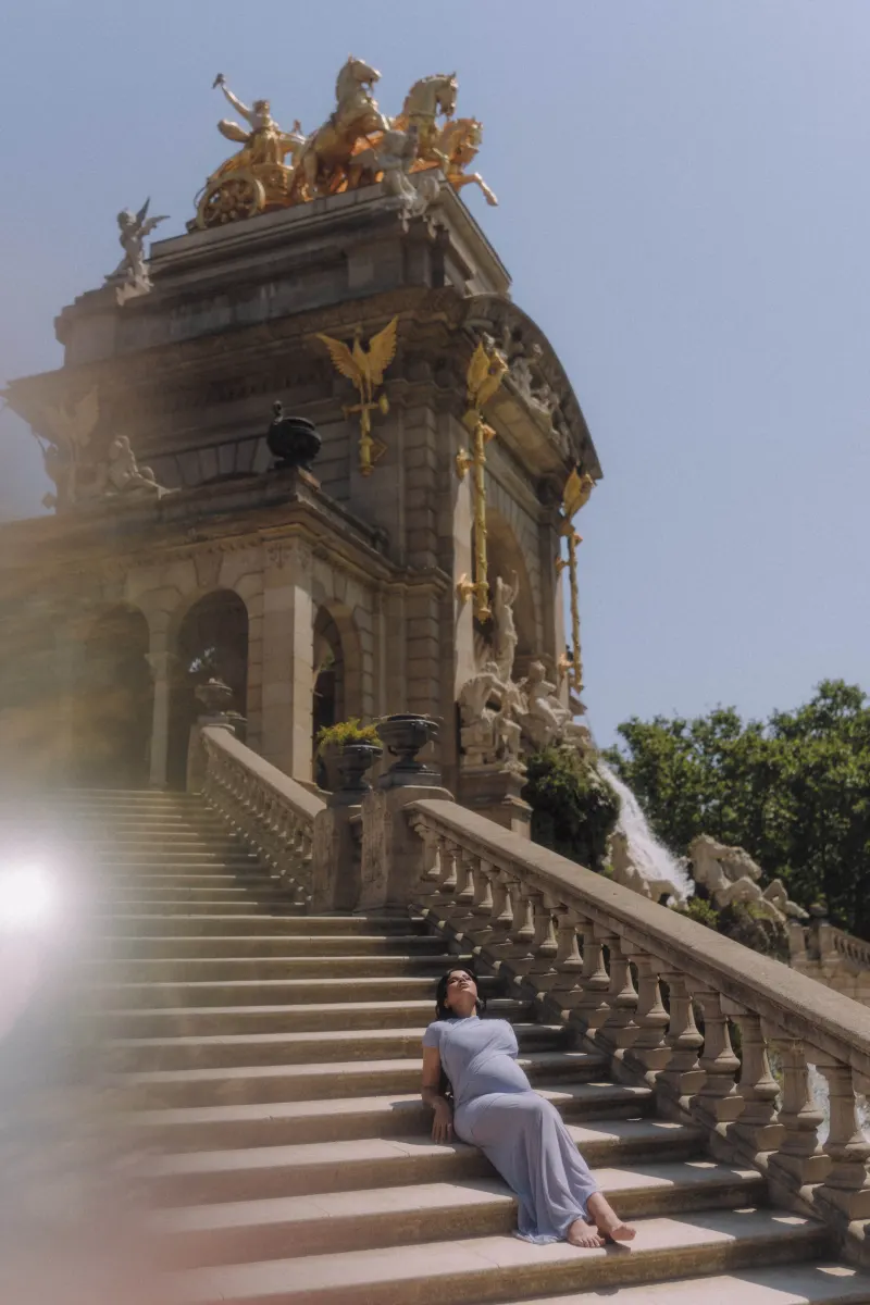 Expectant mother in a pale blue dress relaxes on grand stone steps, Vacation Portraits photoshoot in Barcelona.