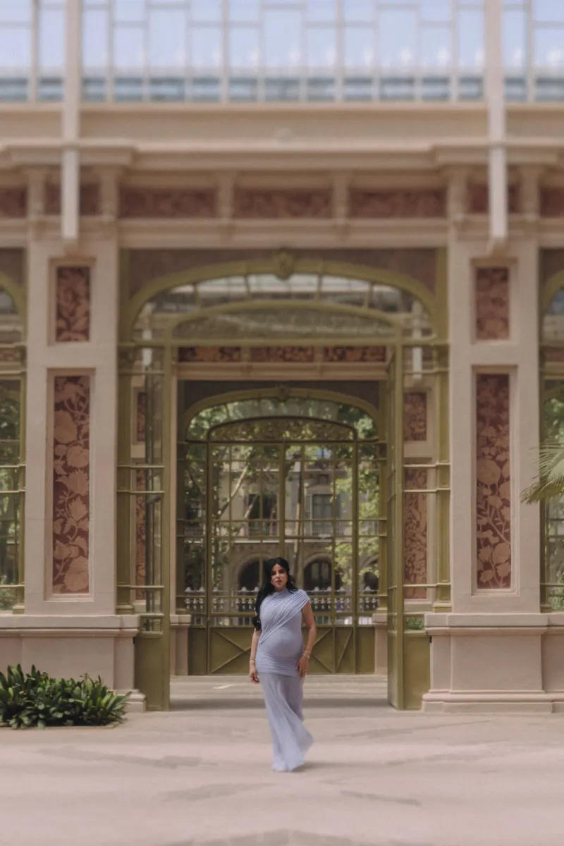 Expectant mother in a pale blue dress stands confidently in a sunlit glass pavilion, Vacation Portraits session.