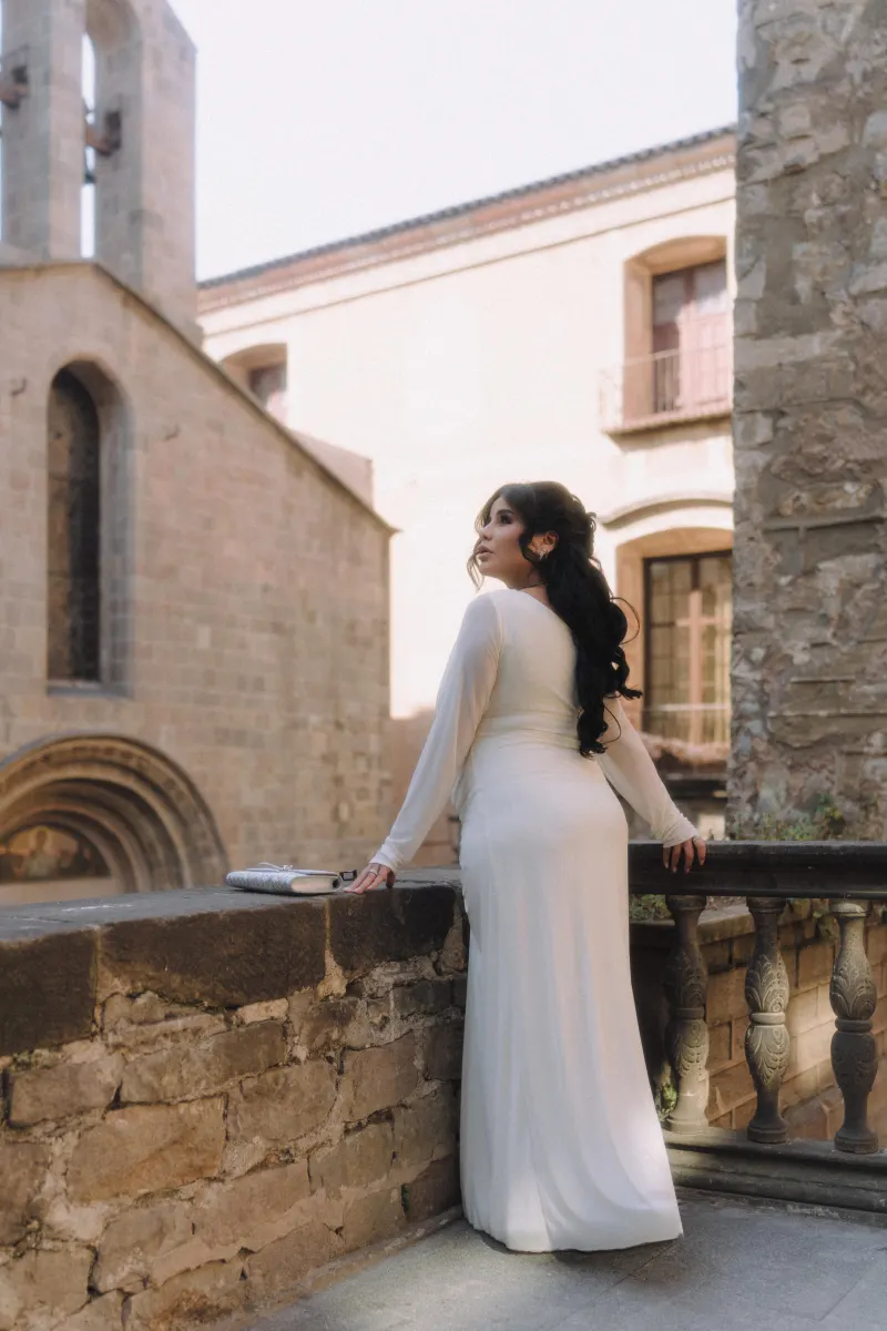 Expectant mother in a white dress gazes upward on a sunlit terrace during a Vacation Portraits photoshoot in a historic court