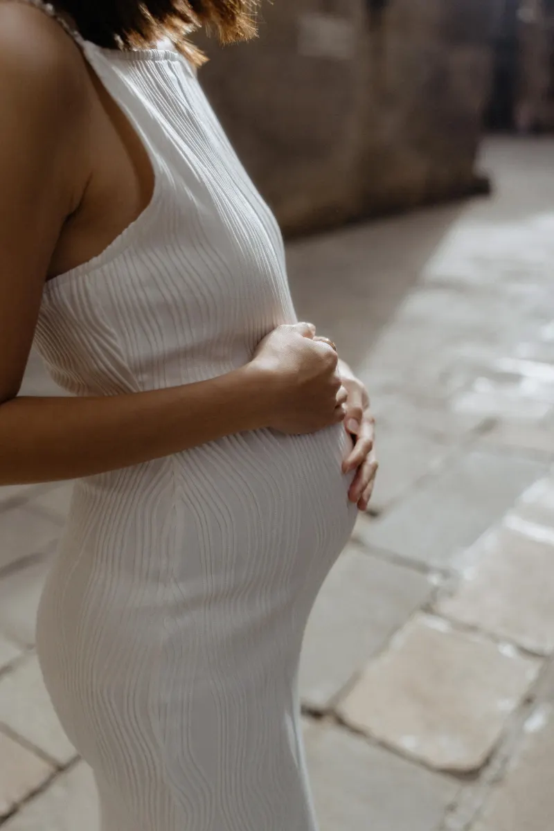 Expectant mother in a white dress gently holding her belly during a Vacation Portraits photoshoot in Barcelona.