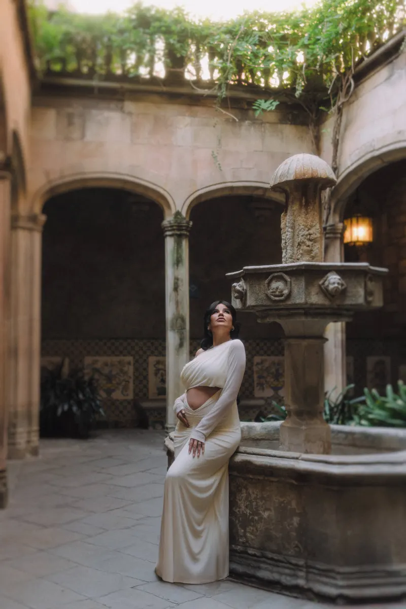 Expectant mother in a white dress leans on a stone fountain, captured during a Vacation Portraits shoot in a tranquil courtya