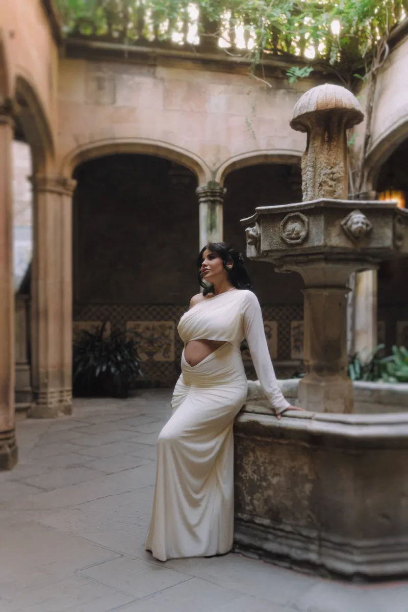 Expectant mother in a white dress leans on a stone fountain during a Vacation Portraits shoot in a peaceful courtyard.