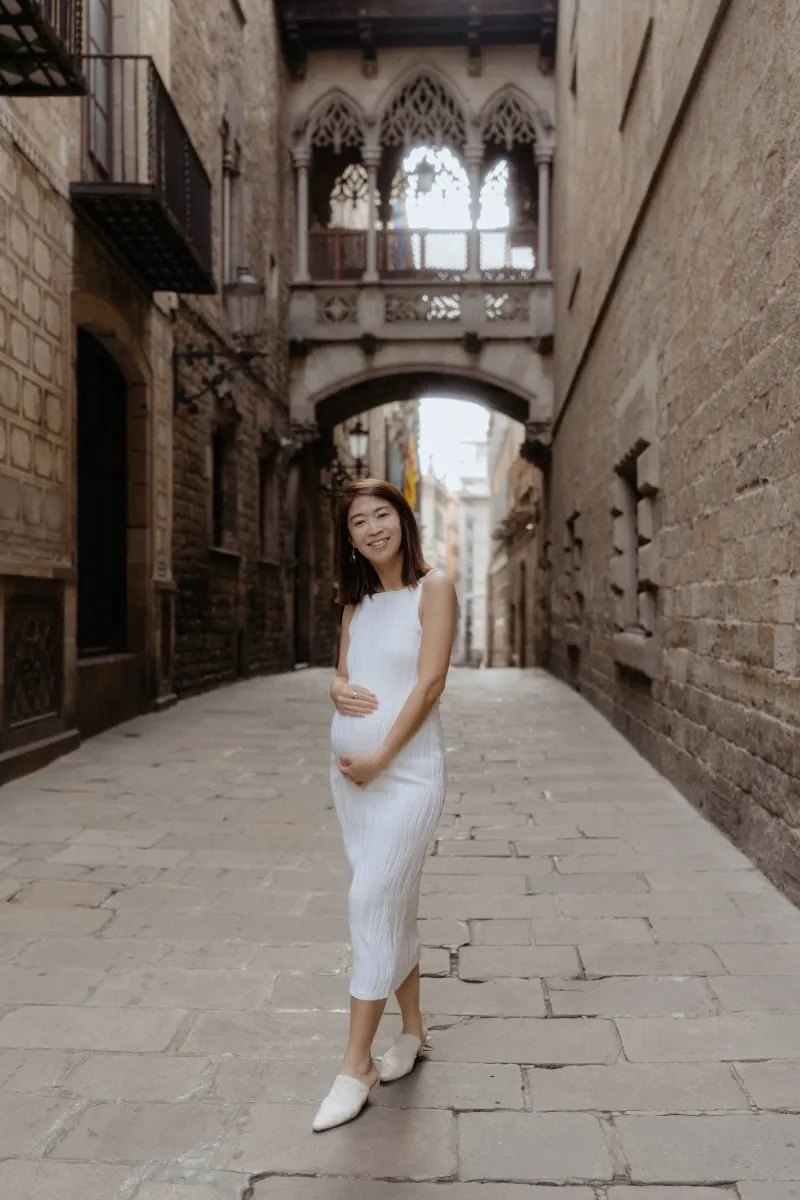 Expectant mother in a white dress smiles warmly during a Vacation Portraits photoshoot in Barcelona’s Gothic Quarter.