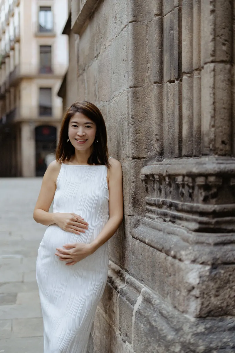 Expectant mother in a white dress smiles while leaning against a stone wall during a Vacation Portraits shoot in Barcelona.