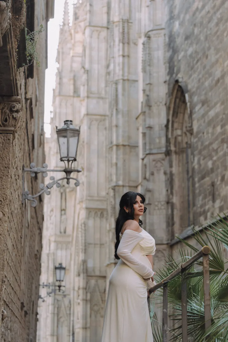 Expectant mother in a white gown gazes thoughtfully in a historic city during a Vacation Portraits photoshoot.