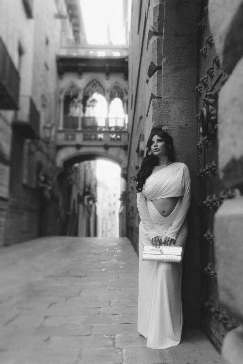 Expectant mother in a white gown leans against a historic stone wall during a Vacation Portraits photoshoot in the city.