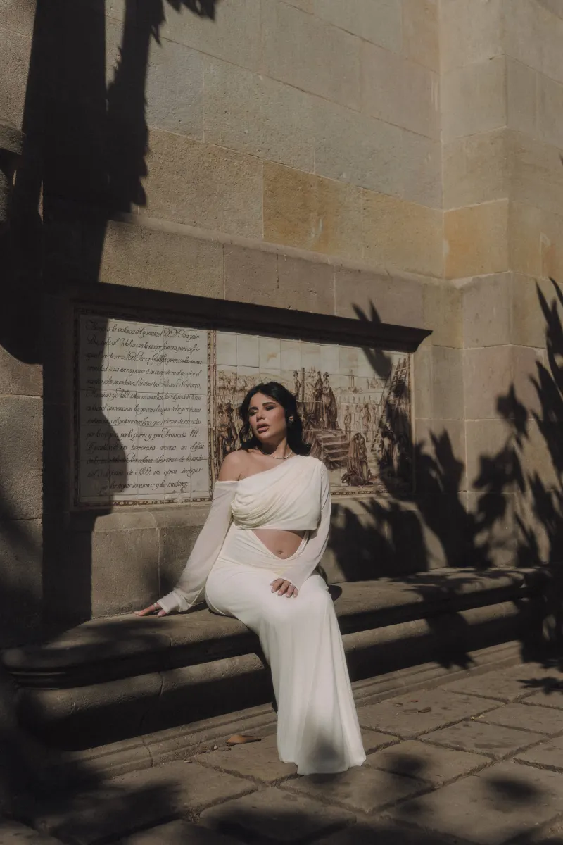 Expectant mother in a white gown rests on a sun-dappled stone bench during a Vacation Portraits photoshoot.