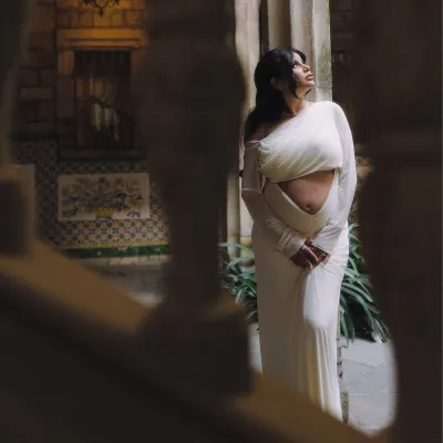 Expectant mother in elegant white dress poses thoughtfully in a historic courtyard during a vacation portraits photoshoot.