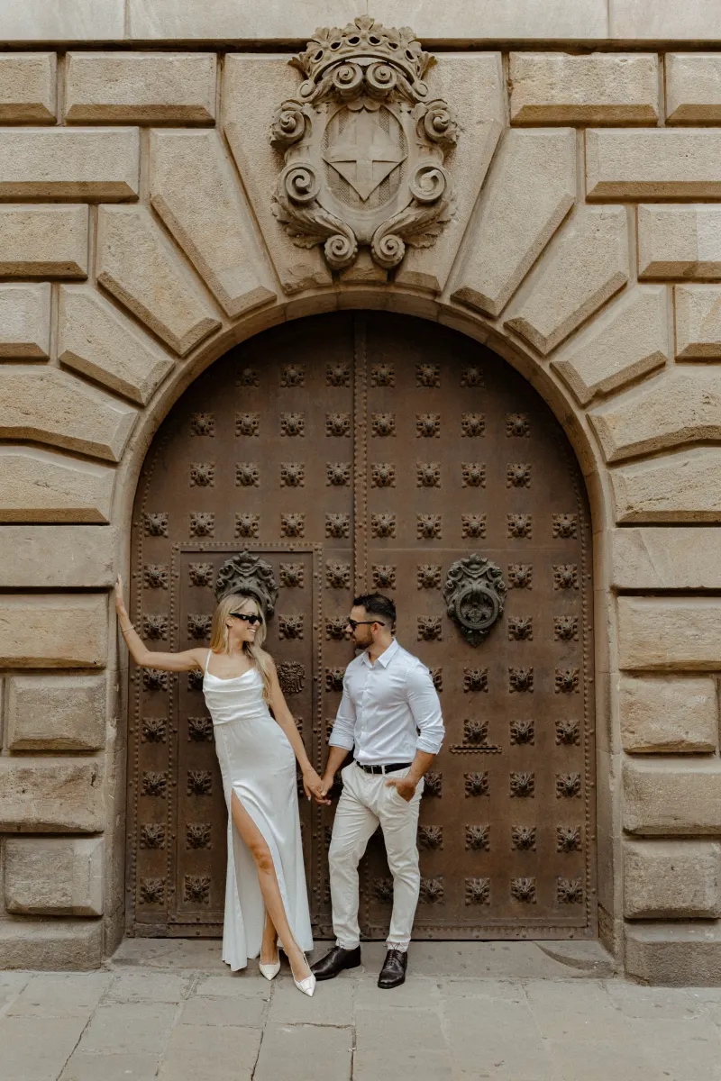 Fashionable couple in white hold hands and share a playful glance by a grand arched doorway during an engagement photoshoot.