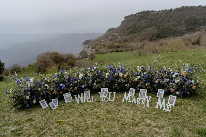 Floral proposal setup on a scenic hillside with “Will You Marry Me” letters and framed couple photos for a surprise shoot.