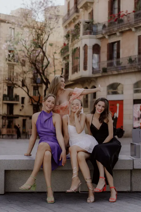 Four friends in stylish dresses share laughter on a city bench, Vacation Portraits photoshoot capturing their joyful bond.