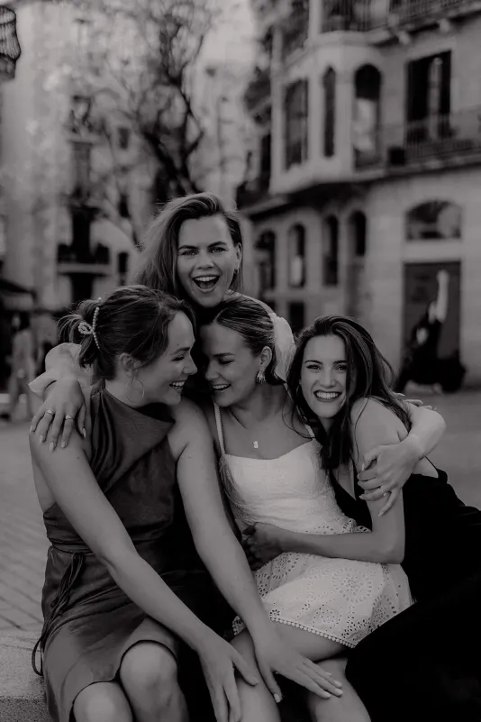 Four women in chic dresses embrace and laugh together on a city street, Vacation Portraits photoshoot capturing friendship.