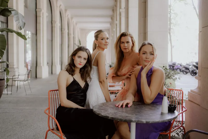 Four women in elegant dresses pose confidently at a city café table, Vacation Portraits photoshoot capturing friendship.