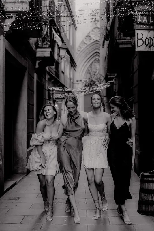 Four women in elegant dresses walk arm in arm, laughing together during a Vacation Portraits photoshoot in a historic city al