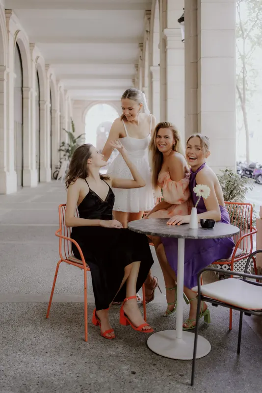 Four women in stylish dresses share laughter at a café table, Vacation Portraits photoshoot in a sunlit city arcade.